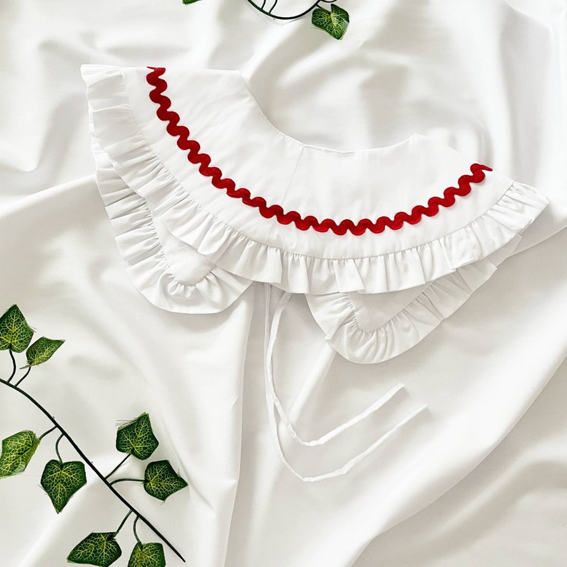 May include: White ruffled collar with a red scalloped trim. The collar is made of a soft, lightweight fabric and has a delicate, feminine look.