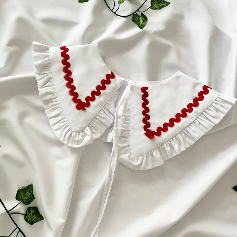 May include: White cotton collar with a red scalloped trim. The collar has a ruffled edge and is designed to be worn as a detachable accessory.