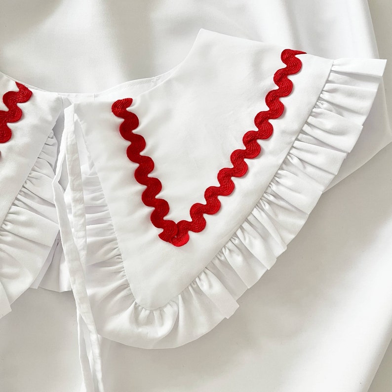 May include: White ruffled collar with a red scalloped trim. The collar has a tie closure.