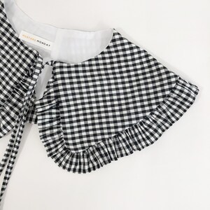 May include: Black and white gingham collar with a ruffled edge and a white lining. The collar has a tie at the back and a label that reads "Mustard Monday".