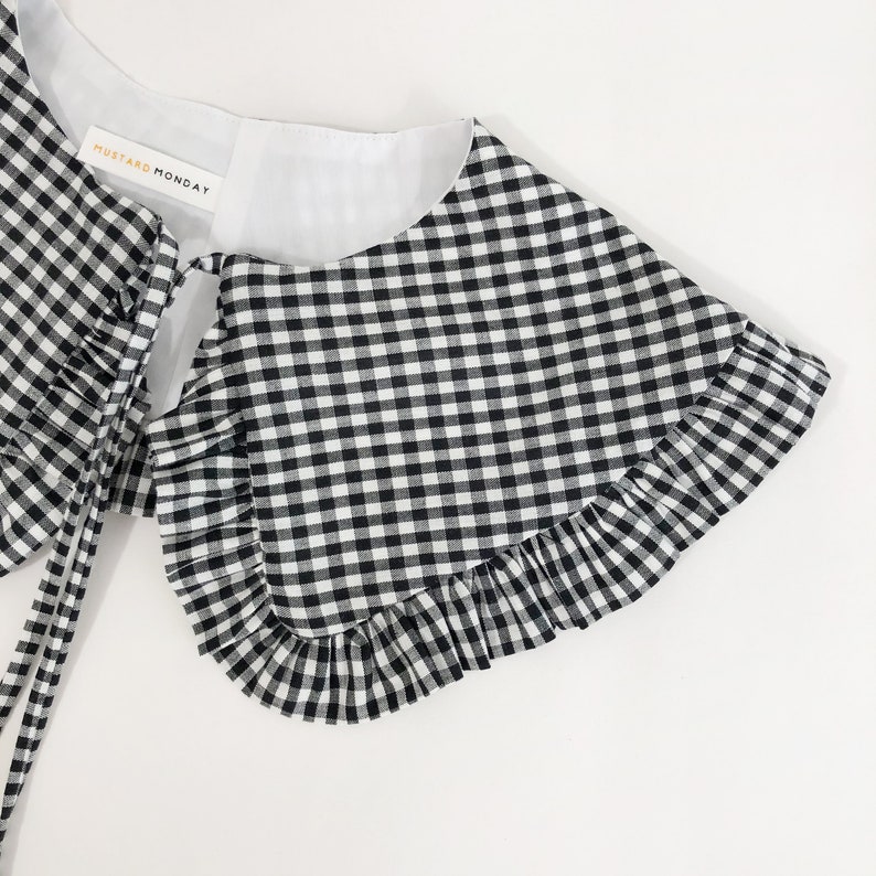 May include: Black and white gingham collar with a ruffled edge and a white lining. The collar has a tie at the back and a label that reads "Mustard Monday".