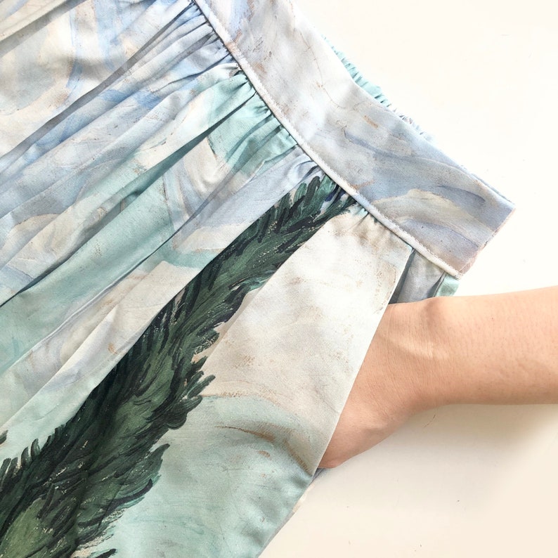 Peut inclure: Jupe bleu clair et blanche avec un imprim&eacute; paysage, avec une main dans la poche. Le tissu pr&eacute;sente un design pliss&eacute; et une impression d&eacute;taill&eacute;e d'arbres et de ciel. Les couleurs de la jupe comprennent des nuances de bleu, de vert et de blanc.