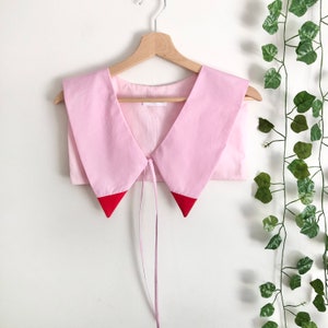 Puede incluir: Un cuello de tela rosa con ribete rojo y cierre de lazo. El cuello está colgado de una percha de madera.