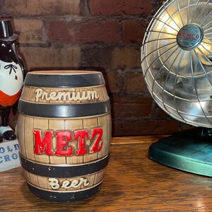 May include: A brown ceramic beer barrel with black bands and the words "Premium Metz Beer" in white and red lettering.