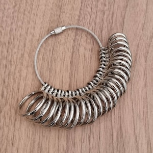 Puede incluir: Una herramienta plateada para medir anillos con múltiples anillos metálicos de diferentes tamaños. Los anillos están unidos a un alambre plateado con un cierre de rosca. La herramienta está diseñada para medir tallas de anillos.