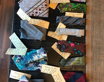 May include: A collection of 14 patterned neckties displayed on a dark wooden surface. The ties are made from recycled materials and feature a variety of colors and patterns, including floral, geometric, and paisley. The ties are labeled with the text "Tie Sticks" and "Up-cycled in Cedar Falls, IA".
