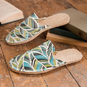 May include: Pair of open-toe slippers with a leaf pattern in shades of green, blue, and black on a white background. The slippers have a natural jute sole and are placed on a wooden surface. Books are in the background.