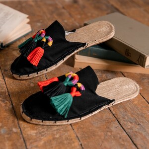 Puede incluir: Sandalias negras sin cordones con detalles de borlas coloridas. Las sandalias tienen una suela de yute tejido y una parte superior de tela negra. Las borlas son rojas, verdes, amarillas y moradas. Las sandalias están sobre una superficie de madera.