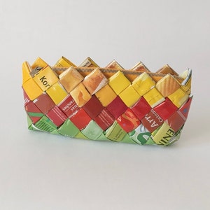 Może przedstawiać: Tkana torebka typu clutch wykonana z recyklingowanych opakowań żywności. Torebka jest żółta, czerwona, zielona i niebieska. Opakowanie zawiera tekst, w tym "Knorr", "Cereal", "Art" i "Enzi".