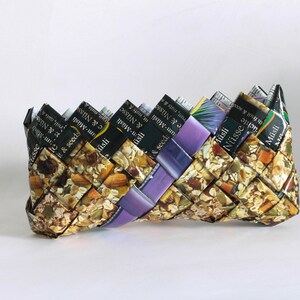 May include: A woven clutch purse made from recycled cereal boxes. The purse is brown and gold with purple accents. The cereal box labels are visible on the outside of the purse. The text on the labels reads "Müsli" and "Nüsse".