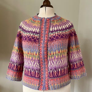May include: A colorful, knitted jacket with a zipper closure. The jacket features a round neckline and three-quarter sleeves. The design incorporates various colors, including purple, pink, yellow, and blue, in a geometric pattern. The jacket is displayed on a mannequin.