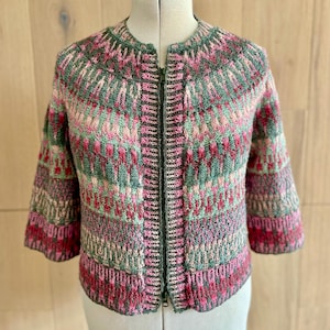 Puede incluir: Chaqueta de punto colorida con cuello redondo y mangas tres cuartos. La chaqueta presenta una cremallera completa y un patrón de tonos rosas, verdes y grises. La prenda se exhibe en un maniquí.