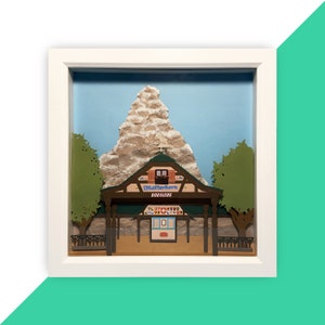 Puede incluir: Un modelo de papel de la atracción Matterhorn Bobsleds de Disneyland. El modelo está enmarcado en un marco blanco y presenta una escultura de papel detallada de la montaña Matterhorn, un pequeño edificio con la inscripción "Matterhorn Bobsleds" en la fachada, y árboles verdes.
