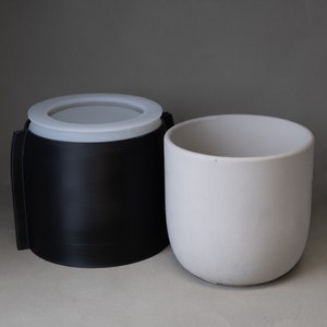 May include: A black container with a white rim next to a small, white, cylindrical pot. The container has a handle-like design on the side. The pot is smooth and has a rounded bottom. The background is a solid gray.