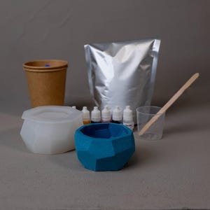 May include: A DIY craft kit with a blue geometric planter, a white geometric mold, a paper cup, small bottles of colored dye, a silver pouch, a clear plastic cup, and a wooden stirring stick. The kit is designed for making decorative items.