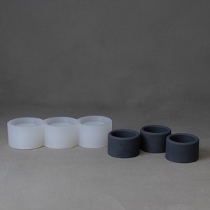 May include: A translucent white silicone mold featuring three connected circular cavities alongside three individual dark gray cylindrical objects with open centers, showcasing a contrast between the flexible mold and the solid finished items.