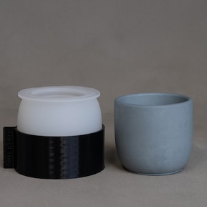 May include: A translucent white silicone mold set inside a black cylindrical plastic frame with the text "Dual Tulip" embossed on the side, positioned next to a solid, smooth, light gray cylindrical cup with a rounded base and open top.