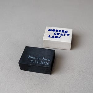 May include: Two square blocks, one white and one dark gray, rest on a light gray surface. The white block has the text "MODERN CRAFT LABS" in blue. The dark gray block is engraved with "Jane & Jack 8.31.2026".