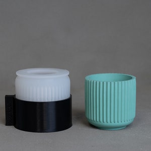 May include: A translucent white cylindrical mold with vertical ridges sits inside a black circular base. Beside it is a finished teal cup featuring a matching vertical ribbed texture on its exterior surface, mirroring the design of the mold.