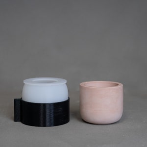 May include: Two small, minimalist planters. One is a light pink, cylindrical pot. The other is a black, rectangular-based container with a white, rounded top. Both are made of a matte material and sit on a neutral gray surface.