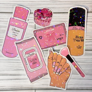 May include: A set of pink and gold holographic stickers featuring makeup and beauty products. The stickers include a bottle of pink mist, a package of facial cleansing wipes, a compact with a bow, a cupcake, a hand holding a makeup brush, and a bottle of foundation with the text "Too Faced Born This Way".