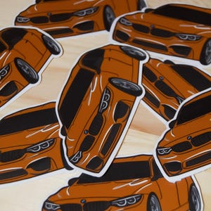May include: A collection of orange car stickers with black detailing. The stickers feature a stylized illustration of a car, with the front grill, headlights, and wheels visible. The stickers are arranged on a light wooden surface.