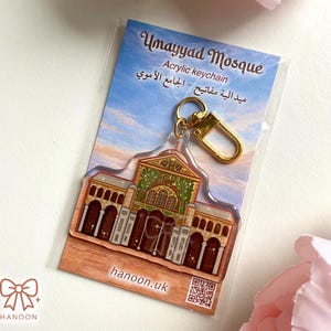 Könnte beinhalten: Ein Acryl-Schlüsselanhänger mit der Umayyaden-Moschee und goldfarbener Hardware. Der Schlüsselanhänger zeigt eine detaillierte Illustration der Moschee mit braunen, grünen und goldenen Akzenten. Die Verpackung enthält den Text "Umayyad Mosque" und "Acrylic keychain".