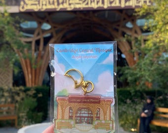 May include: Acrylic keychain featuring the Cambridge Central Mosque. The keychain has a gold-colored clasp and a detailed illustration of the mosque's facade, with benches and a fountain. The background shows the actual mosque building.