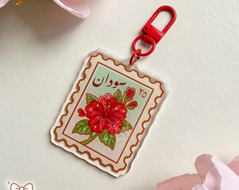 May include: A clear acrylic keychain featuring a red hibiscus flower design, resembling a postage stamp. The stamp has the word "Sudan" in Arabic script and the number 25. It includes a red clasp for attachment.