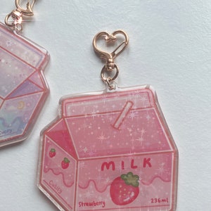 May include: A pink strawberry milk carton-shaped keychain with a gold-colored heart-shaped clasp. The carton reads "MILK" and "Strawberry" with a strawberry graphic. The text "236ml" is also visible. The keychain has a glittery effect.