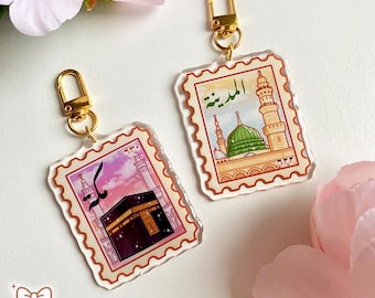 May include: Two clear acrylic keychains with gold-colored hardware. Each keychain features a stamp-like design with religious imagery and Arabic text. One depicts a black structure, the other a green-domed building. The keychains are decorated with pink and white floral accents.