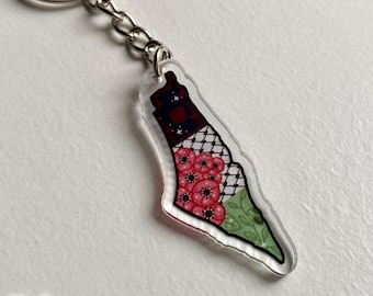 May include: A clear acrylic keychain in the shape of a country. The keychain features a design with red flowers, a checkered pattern, and a green floral pattern. The keychain is attached to a silver metal keyring.