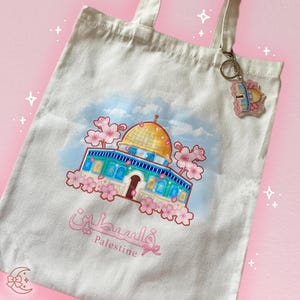 May include: A white canvas tote bag with a colorful illustration of the Dome of the Rock in Jerusalem, surrounded by pink cherry blossoms. The bag also features the Arabic word for Palestine and the English word "Palestine" below the illustration.