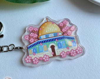 May include: A clear acrylic keychain featuring a detailed illustration of the Dome of the Rock, a famous Islamic landmark in Jerusalem. The dome is depicted in gold with blue and green accents, surrounded by pink cherry blossoms.