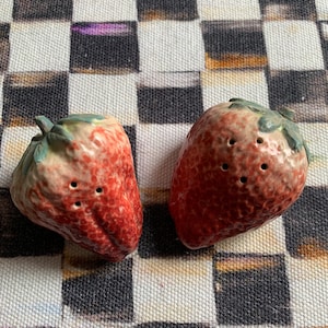 May include: Two red ceramic strawberry salt and pepper shakers with green leaves on a black and white checkered tablecloth.