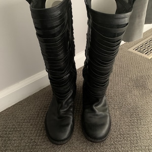 May include: A pair of black leather knee-high boots with a unique design featuring multiple straps that wrap around the leg.