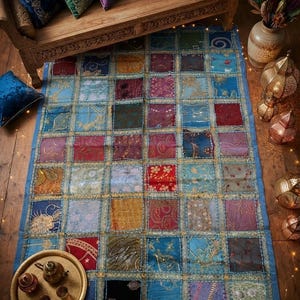 Può includere: Un tappeto patchwork rettangolare caratterizzato da una griglia di quadrati di tessuto multicolore nelle tonalità del blu, rosso, oro e viola. Ogni quadrato è decorato con ricami dorati e paillettes, uniti da cuciture color oro.