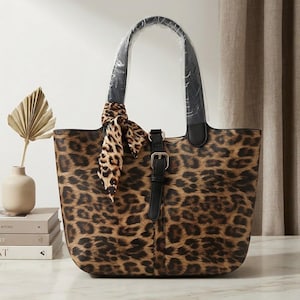 Peut inclure: Un sac cabas avec un motif imprimé léopard marron et noir. Le sac comprend deux anses noires, une sangle verticale centrale noire avec une boucle en métal, et un foulard assorti à motif léopard noué à l'une des anses.