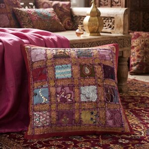 Può includere: Un cuscino quadrato con un design patchwork composto da vari quadrati di tessuto nelle tonalità del rosso, viola, blu e oro. I quadrati sono impreziositi da ricami complessi, paillettes e dettagli in filo metallico, incorniciati da un bordo rosso scuro.
