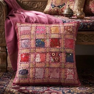 Peut inclure: Un coussin carré présentant un motif patchwork composé de divers carrés de tissu dans des tons de rose, rouge, bleu et or. Les carrés sont séparés par une bordure en treillis brodée dorée et ornés de paillettes, de perles et de broderies florales.
