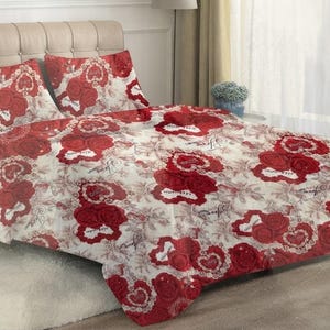 Pode incluir: Um conjunto de roupa de cama com base em tom creme e um padrão repetitivo de rosas vermelho-escuras, motivos de corações em renda branca e texto em cursivo. O conjunto inclui um edredão grande e duas fronhas a condizer.