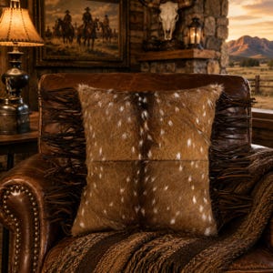 May include: A square throw pillow featuring a brown hide texture with white spots, accented by long dark brown leather fringe along the left and right vertical edges. The pillow has a central horizontal seam and a soft, textured appearance.