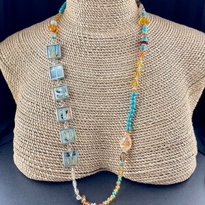 May include: A long necklace with a brown owl charm and a beaded strand with turquoise and amber beads. The necklace also features a silver chain with square pendants that spell out the word "WISDOM".