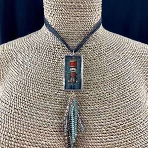 Puede incluir: Un collar de plata con un colgante rectangular de vidrio que presenta una imagen pintada de un soldado de juguete. El colgante está unido a un cordón negro y tiene un borla de cuentas.