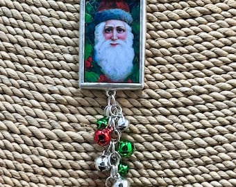 May include: A silver necklace with a rectangular glass pendant featuring a vintage illustration of Santa Claus. The pendant is attached to a chain with a cluster of red and green jingle bells.