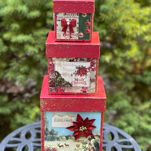 May include: Three red gift boxes with gold glitter stacked on top of each other. The boxes are decorated with Christmas-themed paper and embellishments. The top box has a red bow and the words "Merry Christmas" on it. The middle box has a red poinsettia and the words "Merry Christmas" on it. The bottom box has a winter scene with the words "Have a wonderful Christmas" on it.