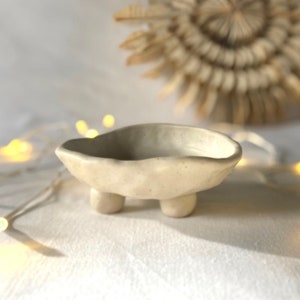 Può includere: Una piccola ciotola in ceramica bianca con una forma irregolare e organica. La ciotola ha tre piccole gambe arrotondate e una superficie leggermente strutturata.