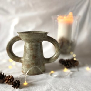 May include: A light green ceramic vase with two handles. The vase has a textured surface and a slightly flared opening. The vase is sitting on a white surface with string lights and pine cones.