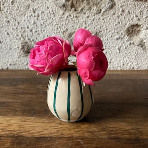 Puede incluir: Un pequeño jarrón de cerámica blanca con rayas verdes contiene rosas rosas. El jarrón está sobre una superficie de madera.
