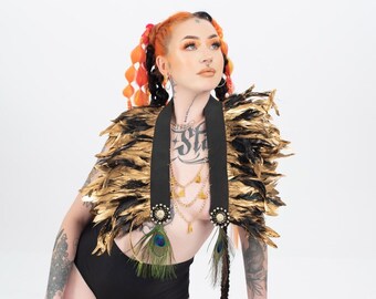 May include: A woman with orange and black hair wearing a gold feather and black strap top with peacock feathers. She is wearing black underwear and has tattoos on her arms and legs. She is leaning on a wooden stool.